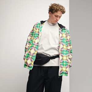 Puede incluir: Una persona que lleva una camiseta blanca, pantalones negros y una chaqueta estampada de color amarillo claro, verde y rosa con una riñonera negra.