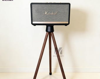 May include: A black and gray Marshall speaker with gold accents, mounted on a dark wood tripod stand. The speaker has the Marshall logo in gold. The stand has three dark wood legs with black rubber feet.