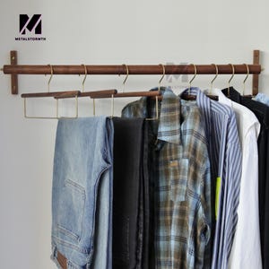 以下が含まれることがあります： 真鍮製のハンガーと様々な衣服を備えた木製の洋服ラック。ラックは壁に取り付けられており、青いジーンズ、黒いパンツ、チェック柄のシャツ、その他のシャツを収納しています。「METALSTORMTH」のロゴが左上に表示されています。