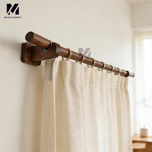 Könnte beinhalten: Ein Gardinenstangen-Set aus Holz mit dunkelbraunem Finish, mit einer zylindrischen Stange und quadratischen Montagehalterungen. Die Stange wird von messingfarbenen Ringen gehalten, und die Vorhänge sind hellbeige.