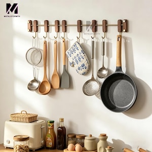 May include: A kitchen wall-mounted rack with various cooking utensils, including a frying pan, wooden spoons, and a floral-patterned oven mitt. The rack is made of wood and metal, and the utensils are hanging from hooks.