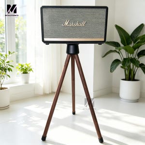 May include: A black and gold Marshall speaker with a mesh front, mounted on a dark wood tripod stand. The speaker has the Marshall logo in gold script. The stand has three legs with black rubber feet. The speaker is in a bright room with plants.