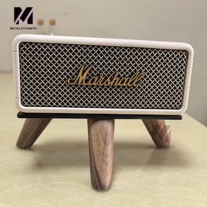 May include: Cream-colored Marshall speaker with a black mesh grill and gold lettering, mounted on a three-legged wooden stand. The speaker has a retro design and the brand name is prominently displayed.