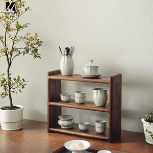 May include: A wooden shelf unit with three shelves, holding a variety of white ceramic teacups, a teapot, and a vase with utensils. The shelf is made of dark wood and has a simple, elegant design.