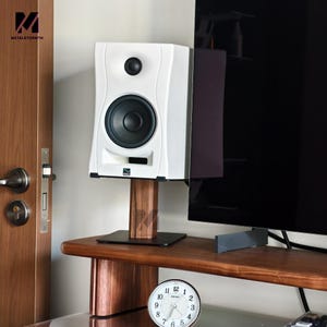 May include: A white speaker with a black speaker cone and a black top button is mounted on a wooden stand. The speaker is positioned near a dark-colored TV screen. A clock with a white face and black numbers is visible.