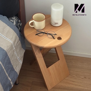 May include: A wooden folding stool with a round top and a hole in the center. The stool is light brown and has a natural wood finish. A white ceramic mug, a pair of eyeglasses, and a white diffuser are on top of the stool.