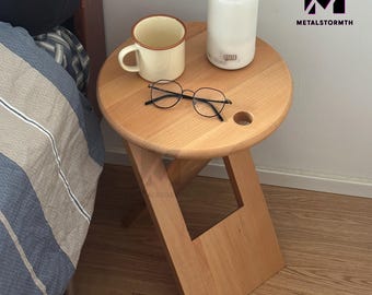 Könnte beinhalten: Ein klappbarer Holzhocker mit runder Oberseite und einem Loch in der Mitte. Der Hocker ist hellbraun und hat eine natürliche Holzoptik. Auf dem Hocker stehen eine weiße Keramiktasse, eine Brille und ein weißer Diffusor.