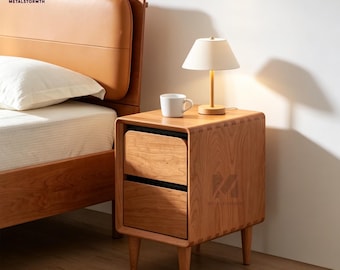 Pode incluir: Uma mesa de cabeceira de madeira com duas gavetas, uma pequena lâmpada e uma caneca branca. A mesa de cabeceira é feita de madeira clara e tem um design arredondado. A lâmpada tem uma abajur bege. A estrutura da cama é visível no fundo.