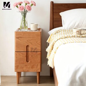 May include: A light brown wooden bedside table with two drawers and a curved handle. The table has four legs. A clear vase with pink flowers and a mug with a bear design are on top. The bed has a white duvet and a yellow blanket with tassels.
