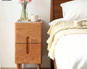 Pode incluir: Uma mesa de cabeceira de madeira clara com duas gavetas e uma pega curva. A mesa tem quatro pernas. Um vaso transparente com flores cor-de-rosa e uma caneca com um urso estão em cima. A cama tem um edredão branco e uma manta amarela.
