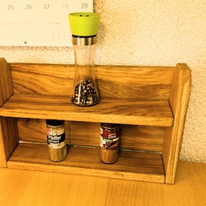 Peut inclure: Un porte-épices en bois à deux étages. L'étagère supérieure contient un moulin à poivre vert avec des grains de poivre noirs et blancs. L'étagère inférieure contient deux pots à épices, l'un étiqueté "Pfeffer Schwarz" et l'autre "Zimt".