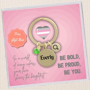 May include: A gold-colored keychain with a heart charm featuring a pink, white, and gray striped design. The keychain also includes a charm with the name "Everly" and a charm with a magnifying glass symbol. The keychain is displayed in a pink gift box with the text "Free Gift Box" and the phrase "In a world of many colors, your love shines the brightest. Be bold, be proud, be you."