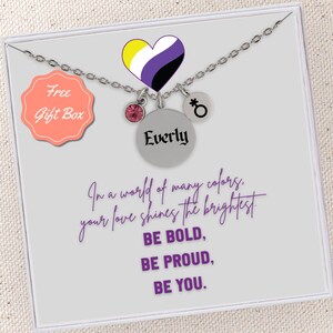 May include: A silver necklace with a purple, yellow, and black heart charm, a pink crystal charm, and a charm with a gender symbol. The necklace is in a white box with the text "Free Gift Box" and "Everly" on the charm. The text "In a world of many colors, your love shines the brightest. BE BOLD, BE PROUD, BE YOU." is printed on the box.