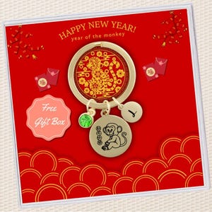 May include: A gold-colored keychain with a red and gold monkey charm, a green crystal charm, and a gold charm with the letter "L". The keychain is in a red box with gold Chinese characters and the text "HAPPY NEW YEAR! year of the monkey".