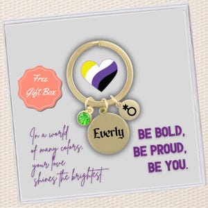 May include: Gold keychain with a heart charm featuring the non-binary pride flag, a green crystal charm, a charm with the gender symbol, and a round charm with the name "Everly" engraved on it. The keychain is in a gift box with the text "Free Gift Box" and the message "In a world of many colors, your love shines the brightest. Be bold, be proud, be you."