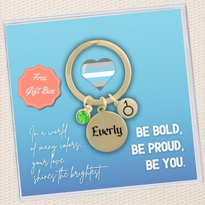 May include: Gold-tone keychain with a heart-shaped charm featuring the transgender pride flag, a green gemstone charm, a charm with the word "Everly" and a charm with a gender symbol. The keychain is displayed in a box with the text "In a world of many colors, your love shines the brightest. Be bold, be proud, be you."