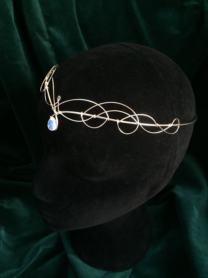 Op de afbeelding: Zilveren draadkroon met een blauwe edelsteenhanger. De kroon bevindt zich op een zwart mannequinhoofd.