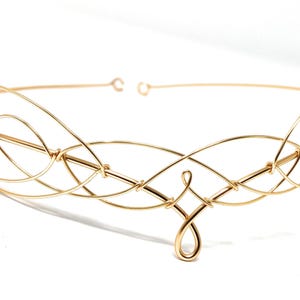 Puede incluir: Una diadema de alambre dorado con un diseño delicado e intrincado. La diadema está hecha de alambre fino que se retuerce y se hace un bucle para crear un patrón fluido y orgánico.