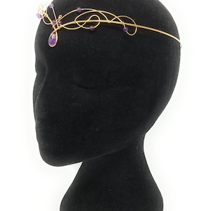 Pode incluir: Uma faixa de cabelo de fio de ouro com uma gema roxa e pequenas contas roxas. A faixa de cabelo está em uma cabeça de manequim preta.