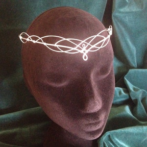 Puede incluir: Una diadema de alambre blanco con un dise&ntilde;o delicado e entrelazado. La diadema est&aacute; en una cabeza de maniqu&iacute; marr&oacute;n oscuro.