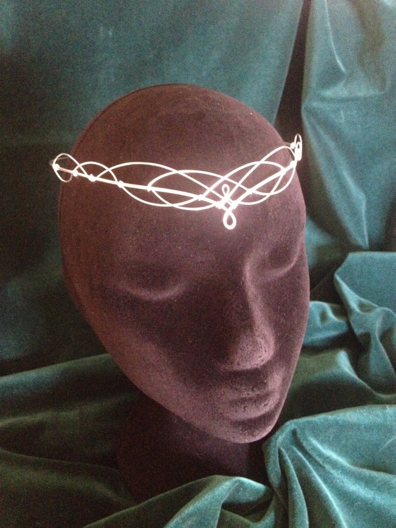 Puede incluir: Una diadema de alambre blanco con un dise&ntilde;o delicado e entrelazado. La diadema est&aacute; en una cabeza de maniqu&iacute; marr&oacute;n oscuro.