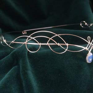 Op de afbeelding: Een zilveren draad hoofdband met een delicaat, verweven ontwerp. Een enkele blauwe edelsteen hangt in het midden.
