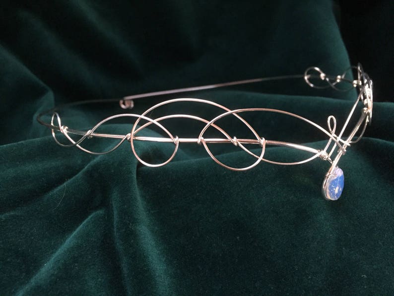 Op de afbeelding: Een zilveren draad hoofdband met een delicaat, verweven ontwerp. Een enkele blauwe edelsteen hangt in het midden.