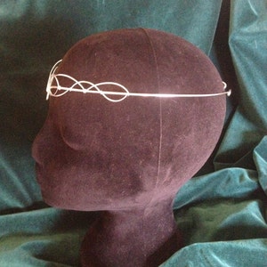 Puede incluir: Diadema de alambre plateado con un dise&ntilde;o delicado e entrelazado. La diadema se muestra en una cabeza de maniqu&iacute; negra sobre un fondo de terciopelo verde.