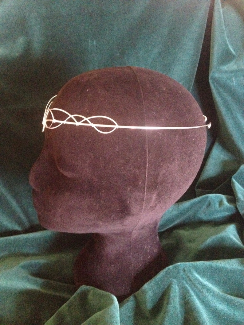 Puede incluir: Diadema de alambre plateado con un dise&ntilde;o delicado e entrelazado. La diadema se muestra en una cabeza de maniqu&iacute; negra sobre un fondo de terciopelo verde.
