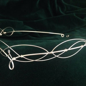 May include: Silver wire crown with a delicate, interwoven design. The crown is made of thin wire that is twisted and looped to create a unique and intricate pattern.
