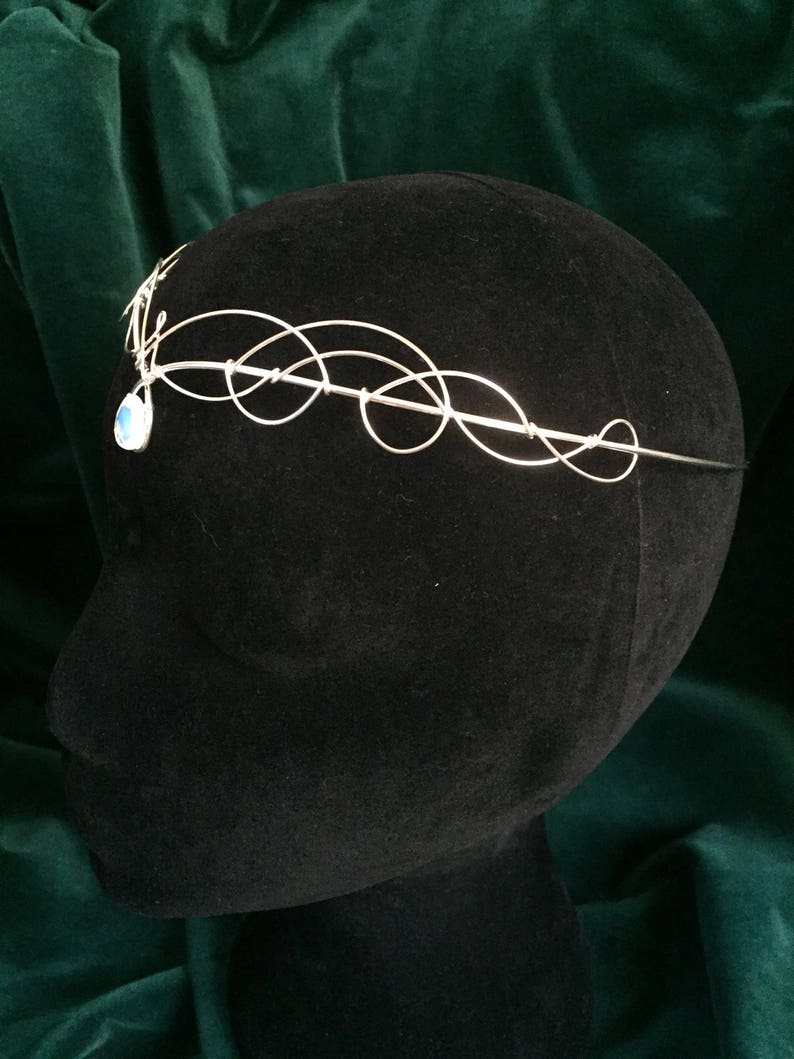 Op de afbeelding: Zilveren draad hoofdband met een delicaat, wervelend ontwerp. Een enkele traanvormige edelsteen hangt aan het midden van het hoofdband.