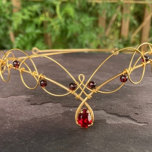 Peut inclure: Une tiare en fil d'or avec une pierre précieuse rouge en forme de larme et de plus petites pierres précieuses rouges le long du fil.