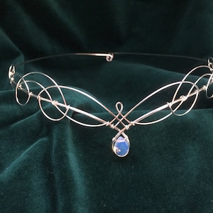 May include: A silver wire headband with a delicate, swirling design. A single blue gemstone dangles from the center.