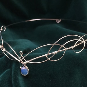 Op de afbeelding: Een zilveren draad hoofdband met een delicaat, verweven ontwerp. Een enkele blauwe opaal steen hangt aan het midden van de hoofdband.