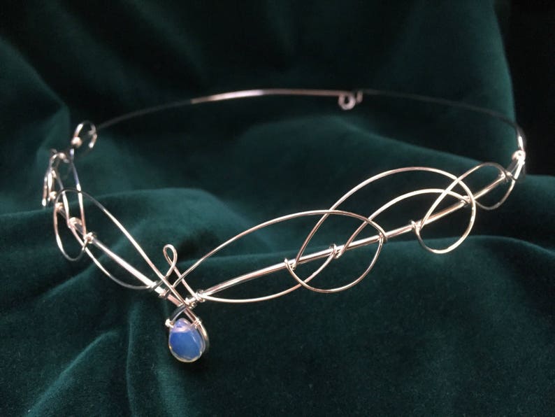 Op de afbeelding: Een zilveren draad hoofdband met een delicaat, verweven ontwerp. Een enkele blauwe opaal steen hangt aan het midden van de hoofdband.