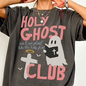 May include: A charcoal gray short-sleeve t-shirt featuring the pink text HOLY GHOST CLUB. The graphic includes a white ghost holding a black book with a cross, a white cross with a gold halo, and small bats with the text ain't no ghost like the holy ghost.