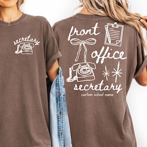 Può includere: T-shirt marrone con testo grafico e illustrazioni bianche. La parte anteriore mostra la parola "secretary" sopra un'illustrazione di telefono. La parte posteriore della maglietta presenta le parole "front office secretary" con illustrazioni di fiocco, blocco note e stelle.
