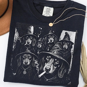 May include: A black t-shirt featuring a graphic print of five dachshunds wearing pointed witch hats. The dogs are depicted with a cigarette in their mouths and wearing pentagram necklaces. The shirt has a Comfort Colors brand label at the neck.