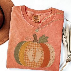 May include: A terracotta-colored short-sleeve t-shirt featuring a large pumpkin applique on the front. The pumpkin is composed of various fabric patches in shades of green, orange, and beige with a central graphic of two baby footprints.