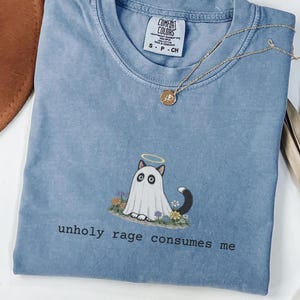 May include: A blue short-sleeved t-shirt featuring a graphic of a cat wearing a white ghost sheet with a yellow halo, surrounded by small flowers. The text "unholy rage consumes me" is printed in black below the graphic. The shirt has a Comfort Colors label.