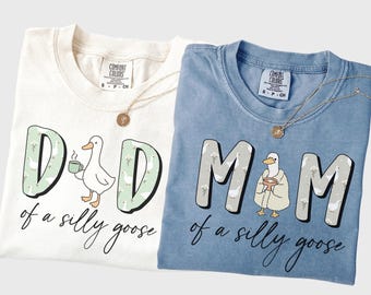 May include: Two Comfort Colors t-shirts, one cream and one blue, with the phrases "Dad of a silly goose" and "Mom of a silly goose" in a whimsical font. Each shirt features a cartoon goose illustration and a gold necklace.