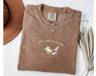 May include: Brown Comfort Colors t-shirt with the phrase "off to fall on my ass" and a cartoon frog wearing ice skates. A silver necklace with a pendant rests on the shirt. A brown hat is visible in the upper left corner.