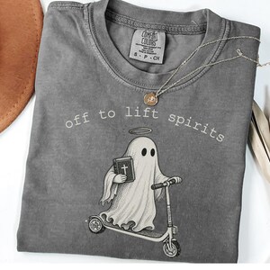 May include: A charcoal gray short-sleeve t-shirt featuring a graphic of a ghost with a halo riding a scooter and holding a book. The text "off to lift spirits" is printed in white above the ghost. The shirt has a Comfort Colors brand label.