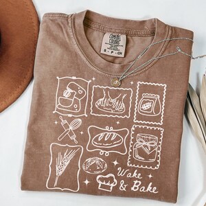 Può includere: Una maglietta marrone Comfort Colors con una grafica bianca di immagini relative alla panificazione e la scritta "Wake & Bake". Una collana d'argento con un ciondolo è drappeggiata sulla maglietta. Un cappello marrone è visibile in alto a sinistra.