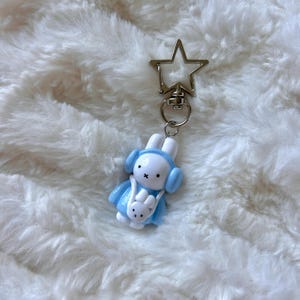 May include: A blue and white bunny keychain with a star-shaped metal clip. The bunny is wearing blue earmuffs and carrying a smaller white bunny.