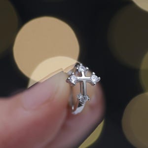 May include: A silver earring featuring a cross design, embellished with small, clear gemstones. The cross is the central element, with the gemstones providing sparkle. The earring is held against a blurred backdrop of warm, out-of-focus lights.