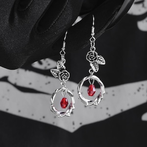 Könnte beinhalten: Silberne Ohrringe mit einem roten Kristall, der an einem runden Design hängt. Die Ohrringe haben ein Rosenmotiv mit Blättern.