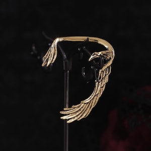 Op de afbeelding: Een gouden oormanchet met een vleugelontwerp. De manchet is gebogen en heeft een delicaat, ingewikkeld ontwerp.