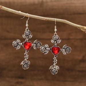 May include: A pair of silver earrings with a cross design and red heart-shaped crystals. The cross is adorned with rose details.