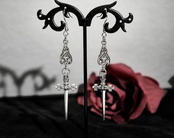 May include: A pair of silver earrings with a sword design. The swords have a detailed cross guard and a pointed tip. The earrings are hanging from a black stand.